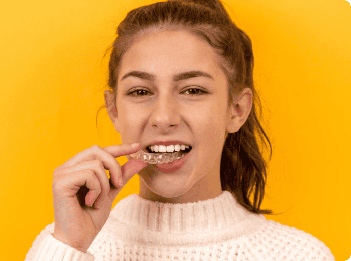 Bright yellow background, with a woman confidently smiling at the camera holding an Invisalign fitting to her teeth.
